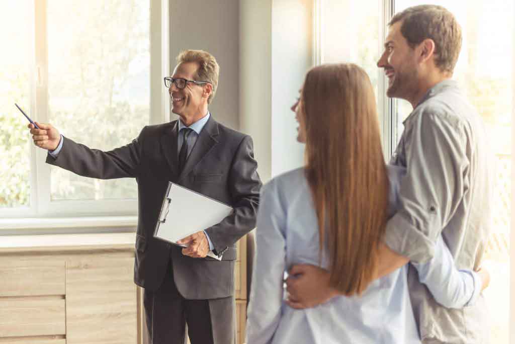 Handsome middle aged realtor in classic suit and eyeglasses is holding a notepad and smiling while showing new apartment to young couple